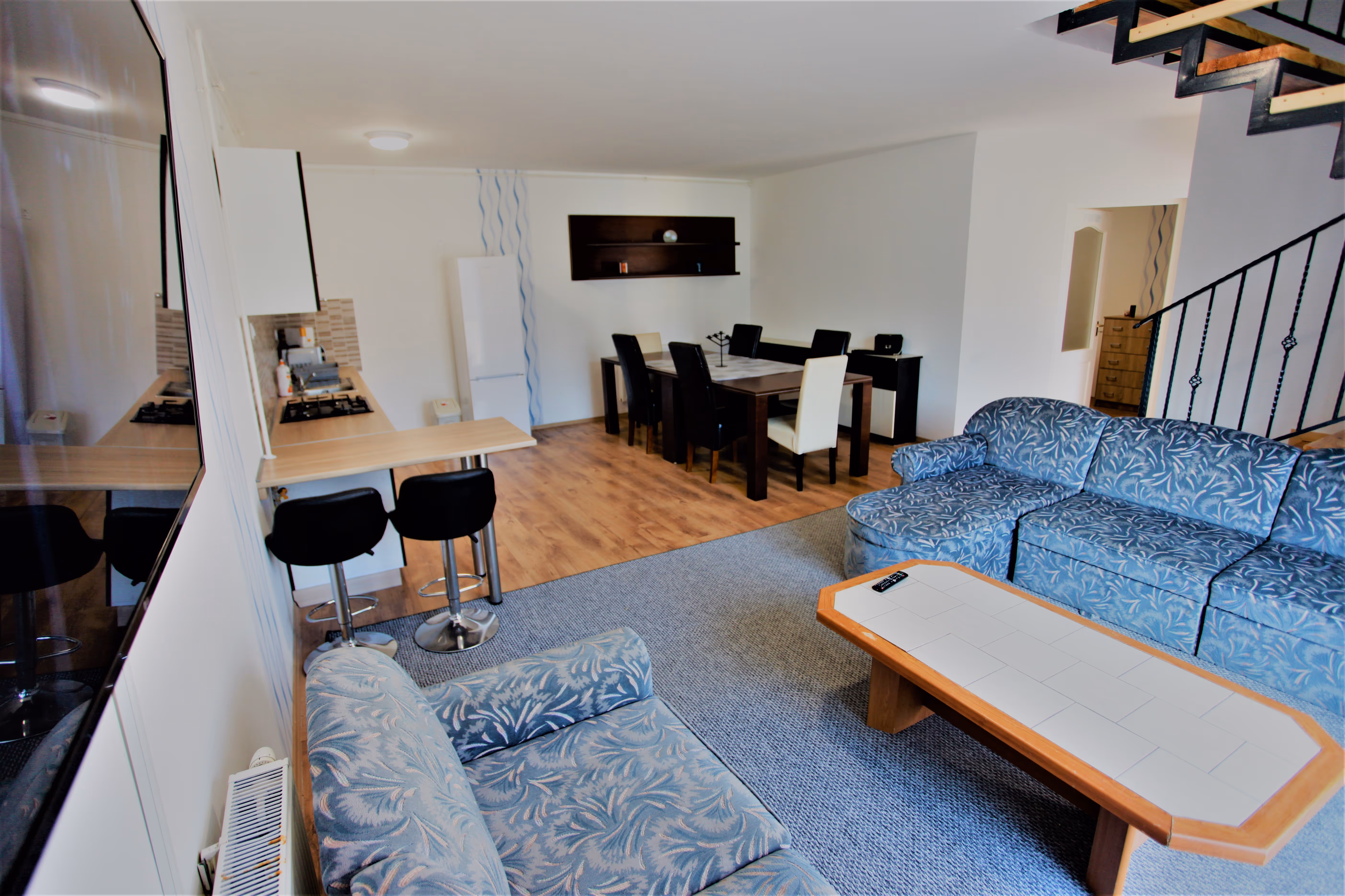 Tágas nappali kék mintás kanapékkal, étkezőasztallal és konyhapulttal bárszékekkel